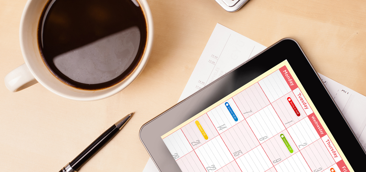 Close up of an office table with a cup of coffee, pen, laptop, and tablet displaying a calendar