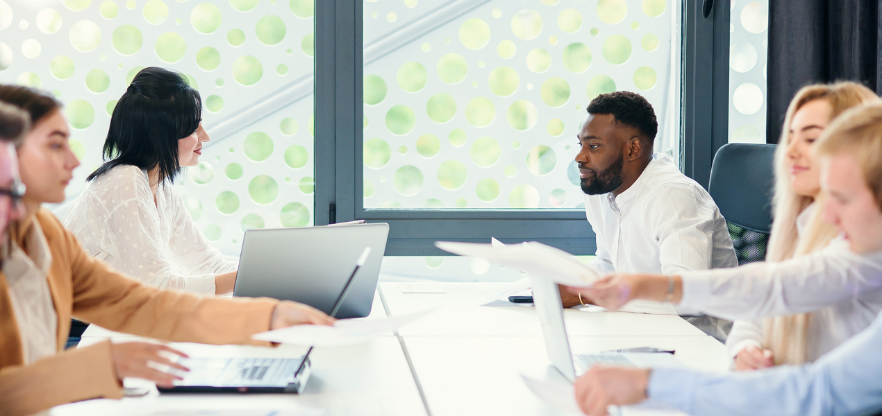 A group of diverse individuals seated at a table, each using a laptop for collaborative work or discussion.