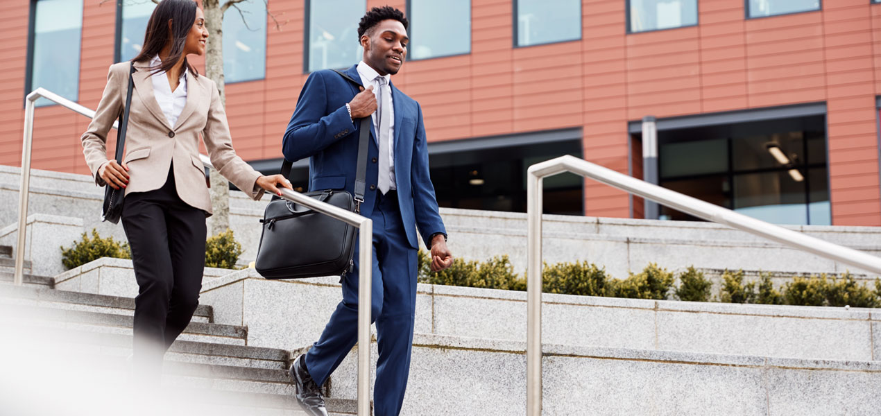 Two business professionals, a man and a woman, are walking down a set of stairs outside.