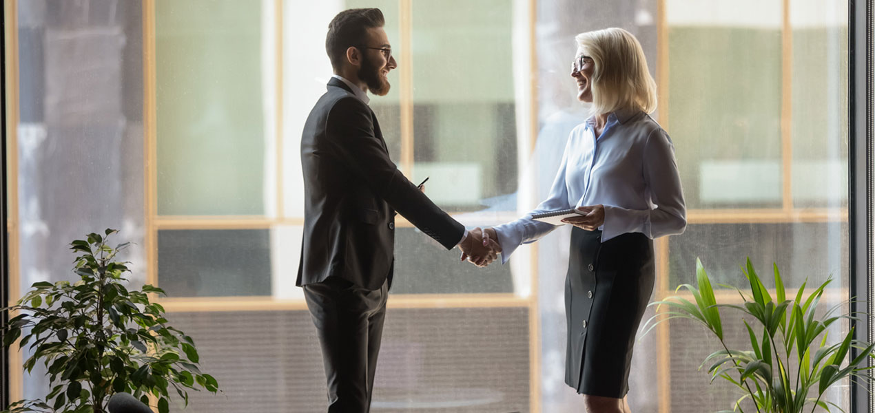 A business man and business woman shaking hands in front of a floor-to-ceiling window.
